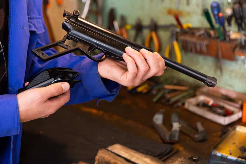 Person holding a firearm with detached grip components in a workshop or gunsmithing environment with tools visible in the background