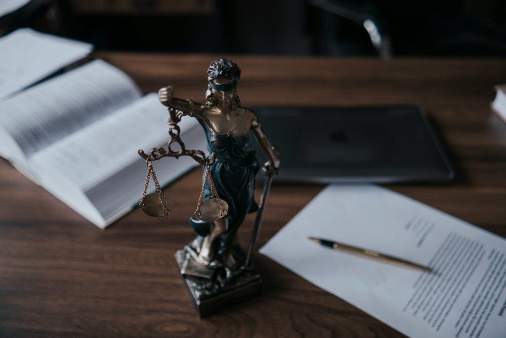 Bronze statue of Lady Justice holding balanced scales and a sword, placed on a wooden desk surrounded by an open law book, printed legal documents, and a pen, with a dark, softly lit office background suggesting legal analysis, fairness, and the judicial process.