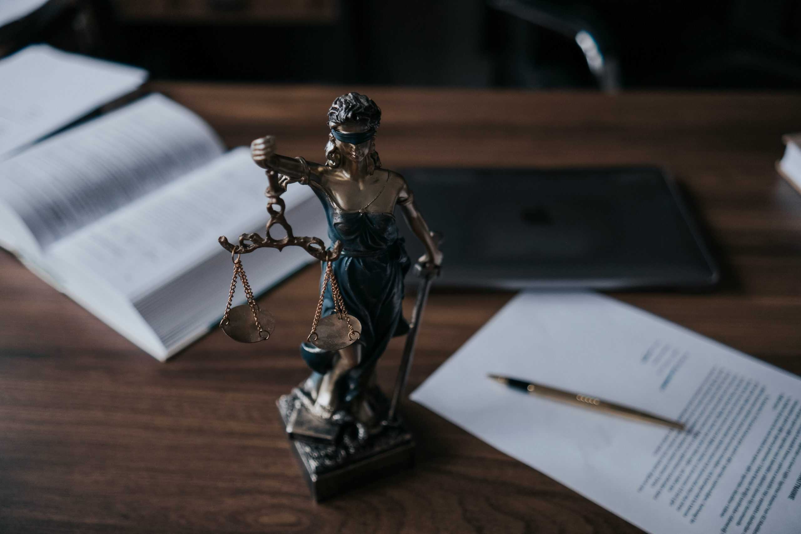 Bronze statue of Lady Justice holding balanced scales and a sword, placed on a wooden desk surrounded by an open law book, printed legal documents, and a pen, with a dark, softly lit office background suggesting legal analysis, fairness, and the judicial process.