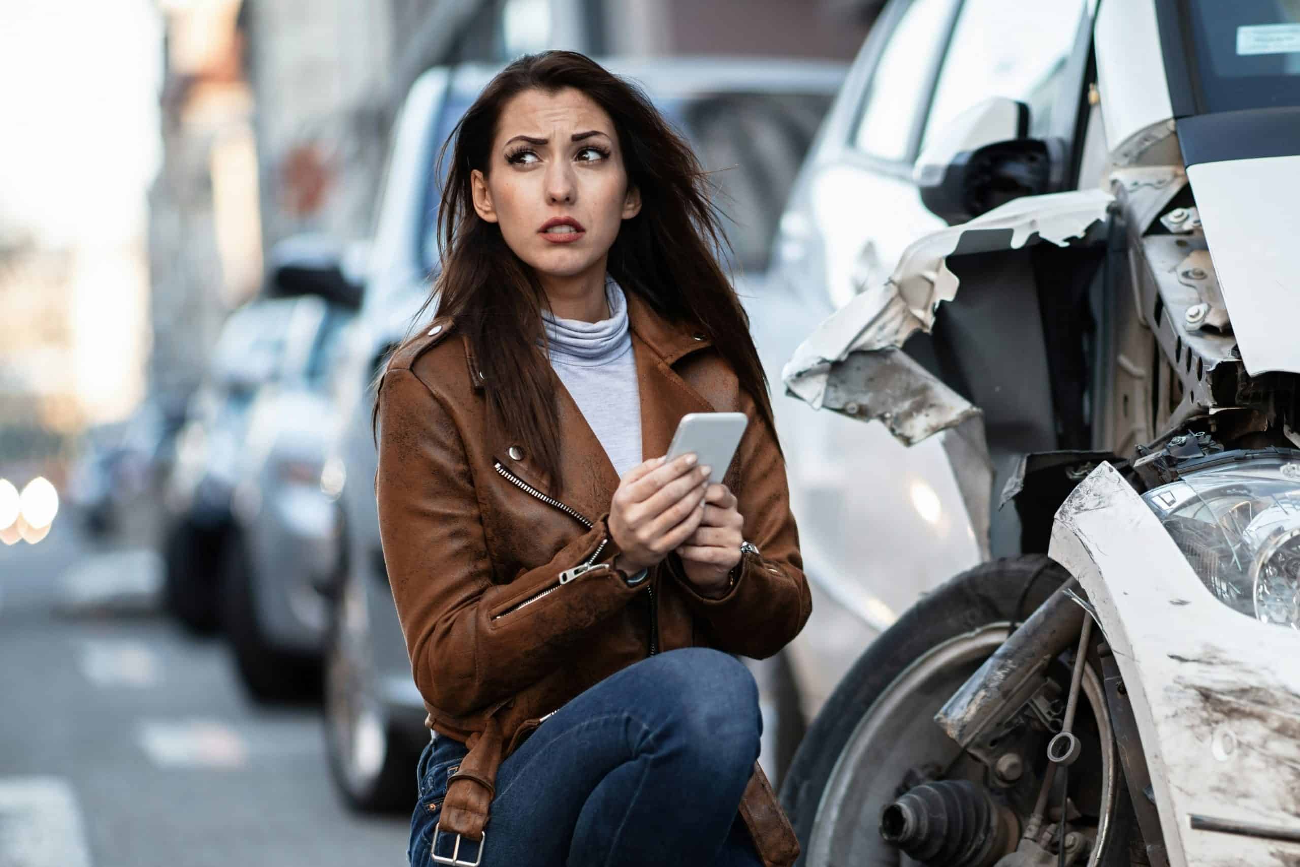 Woman crouching beside a damaged car on a city street, holding a smartphone and looking concerned. The front of the vehicle shows visible crash damage with a broken bumper and exposed components, while other cars are parked along the road in the background. The woman is wearing a brown jacket and jeans, and the scene suggests the aftermath of a traffic accident.