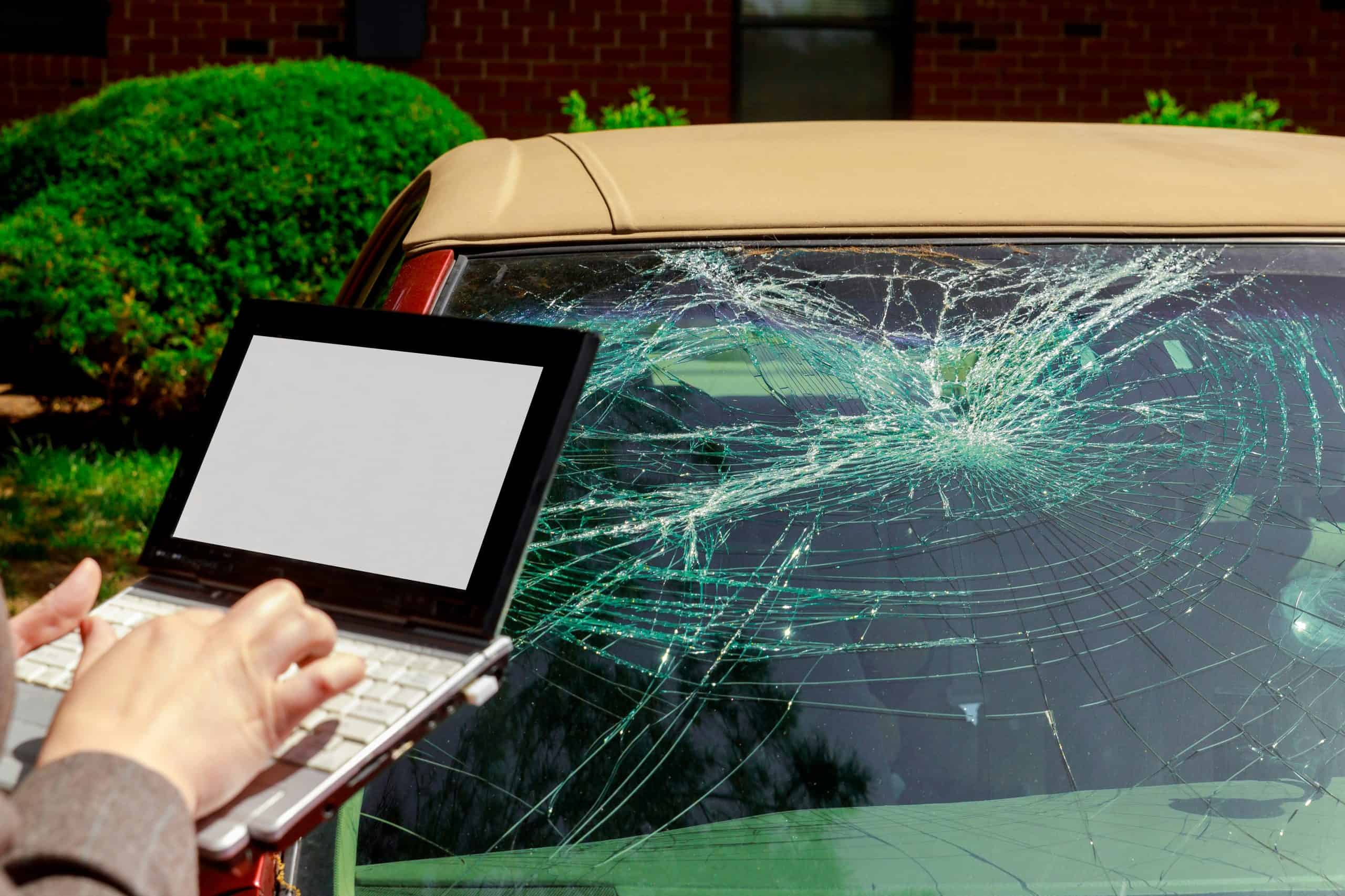 Close-up of a severely cracked car windshield with spiderweb-like fractures spreading across the glass, while a person in the foreground holds a small laptop with a blank white screen and types on the keyboard, suggesting documentation or reporting of vehicle damage, with green bushes and a brick building visible in the background under daylight.