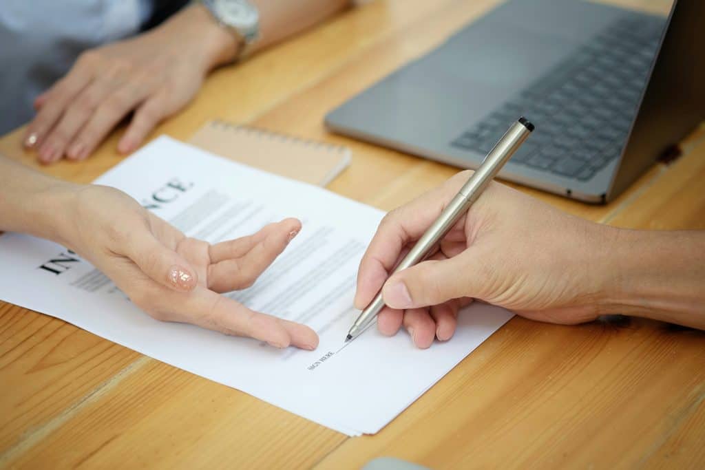 Close-up of two people’s hands over a printed contract on a wooden desk, one person holding a silver pen poised above the signature line while the other gestures toward the document, with a laptop and small notebook blurred in the background, suggesting a formal agreement or document signing in an office setting.