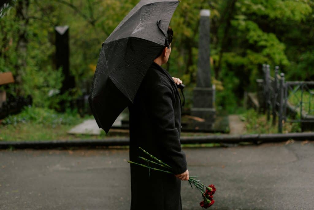 Person dressed in a black coat standing outdoors at a cemetery on a rainy day, holding a black umbrella overhead and a small bouquet of red flowers in one hand, with gravestones, greenery, and a damp pathway visible in the background, creating a somber and reflective atmosphere.