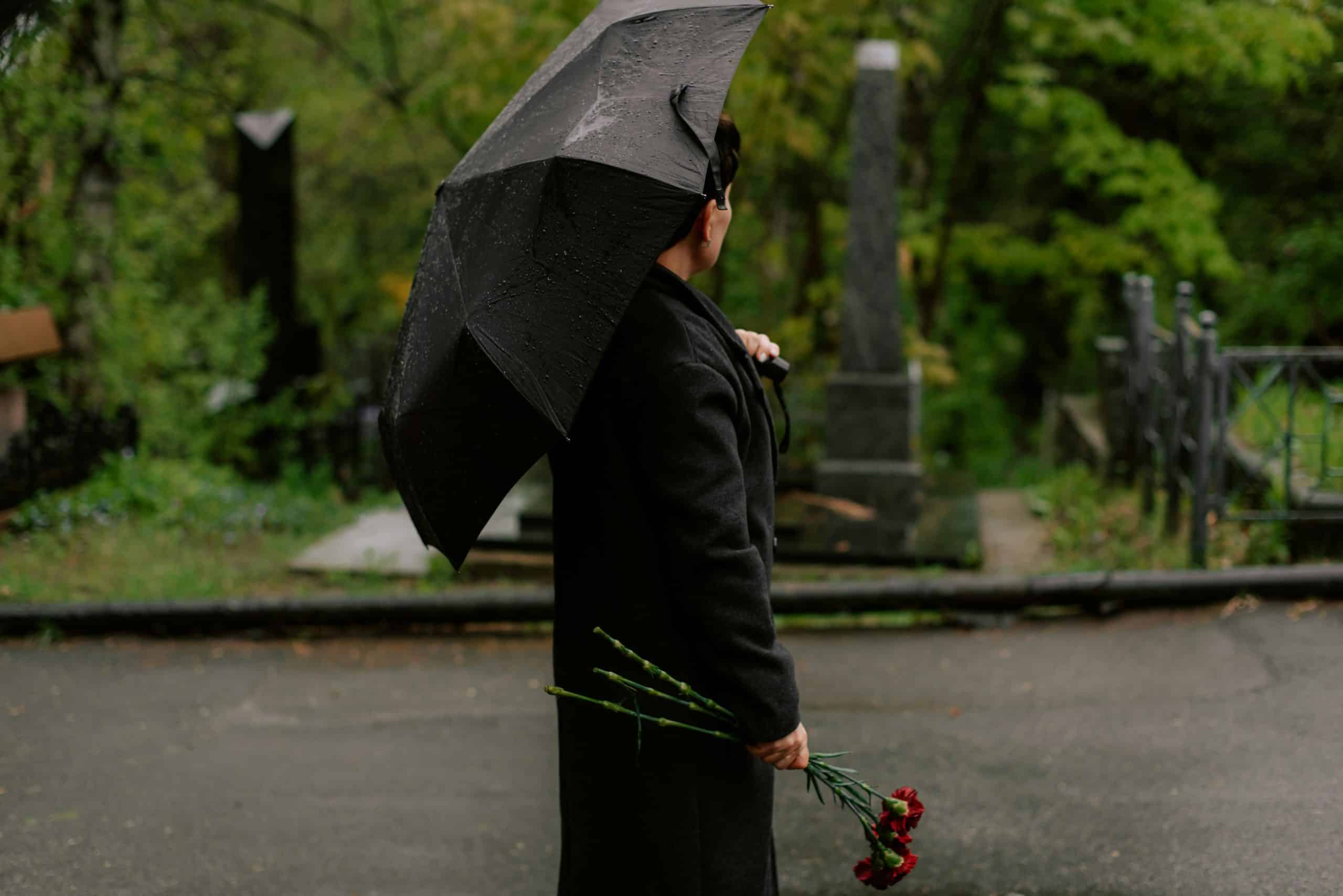 Person dressed in a black coat standing outdoors at a cemetery on a rainy day, holding a black umbrella overhead and a small bouquet of red flowers in one hand, with gravestones, greenery, and a damp pathway visible in the background, creating a somber and reflective atmosphere.