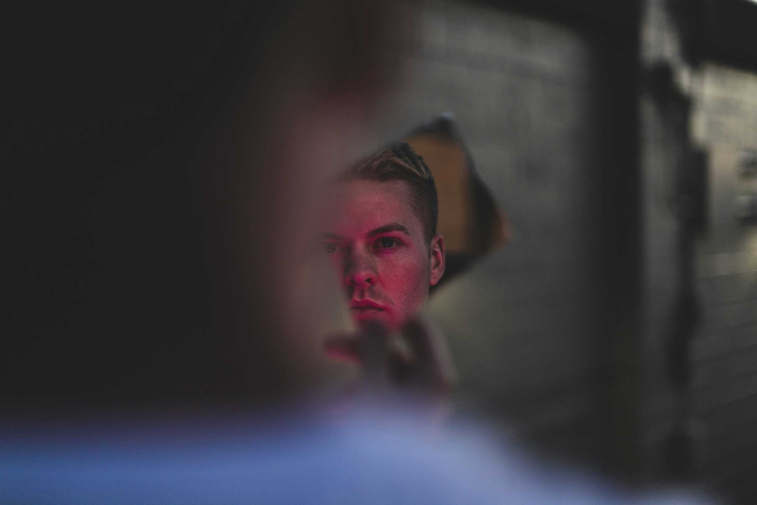 Young man looking at his reflection through a jagged hole in a damaged mirror or wall, his face partially illuminated with red light while the foreground remains blurred, creating a tense and introspective atmosphere.