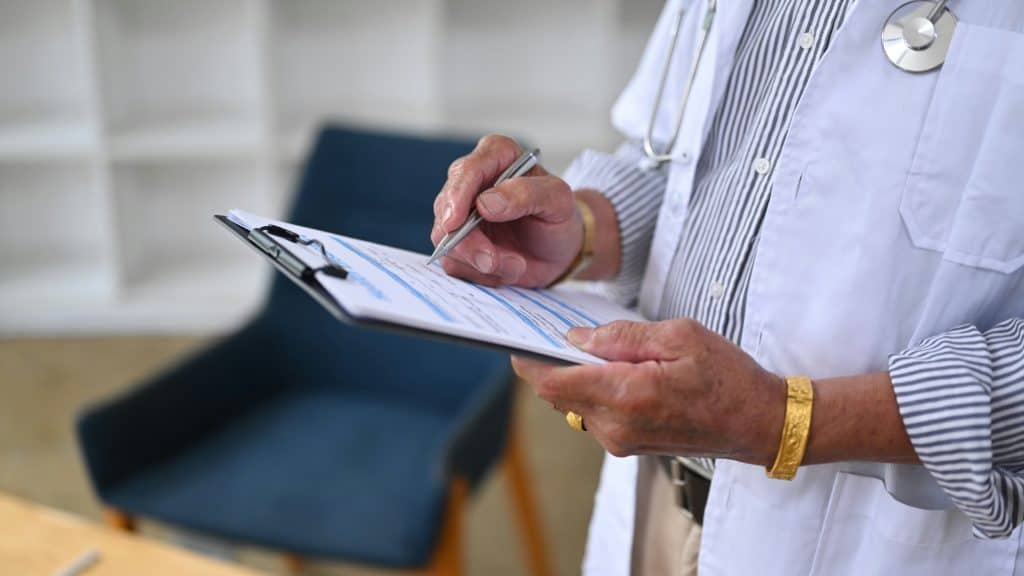 A doctor in a white coat writes notes on a clipboard in a clinical setting.