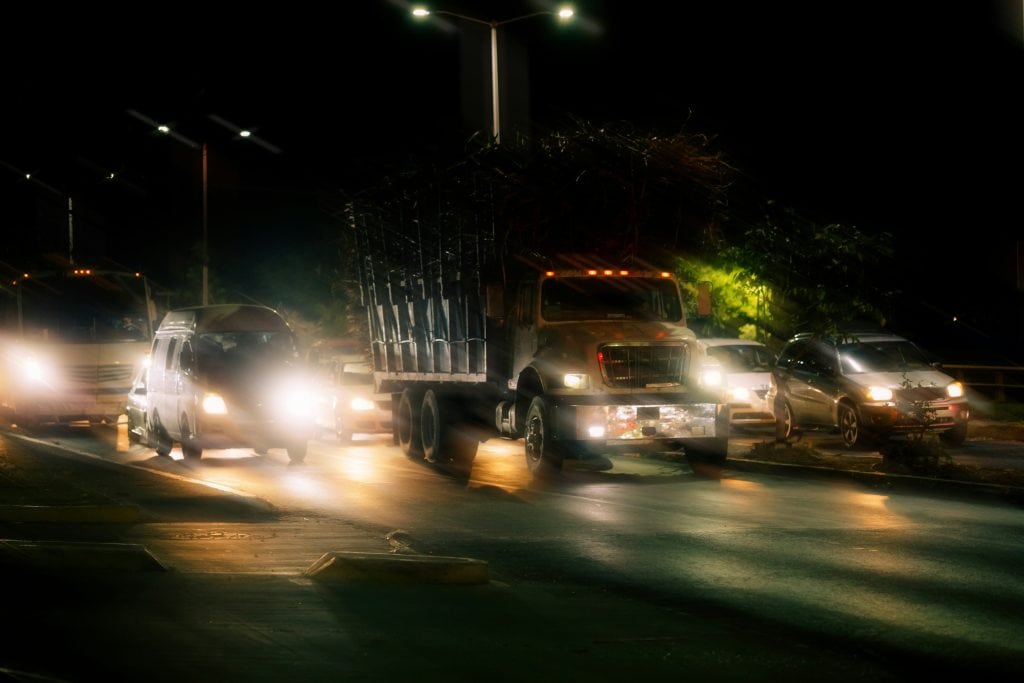 A truck drives down a dimly lit street at night, with headlights illuminating the road ahead.