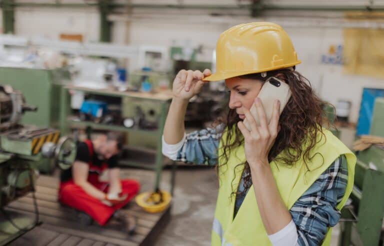 A woman in a hard hat is talking on a cell phone, likely discussing work-related matters on a construction site.