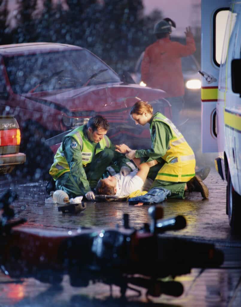 Paramedics providing emergency care to an injured person lying on a wet roadway after a car accident, with damaged vehicles and an ambulance nearby in rainy conditions.