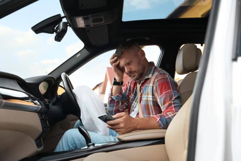 A man sitting in the driver's seat of a car, holding a cell phone and looking at the screen.