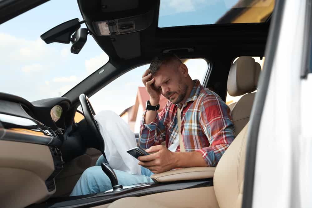 A man sitting in the driver's seat of a car, holding a cell phone and looking at the screen.