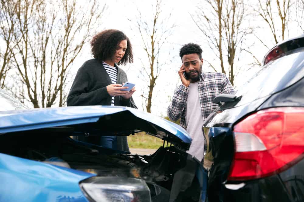 A man and woman talk on the phone beside a damaged car after a car accident.