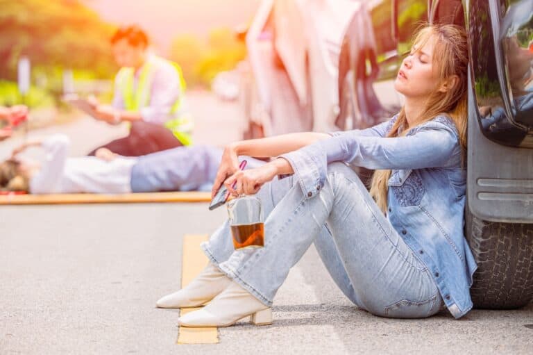 A woman sitting on the roadside, visibly distressed, near a car involved in an accident. Emergency responders are nearby.