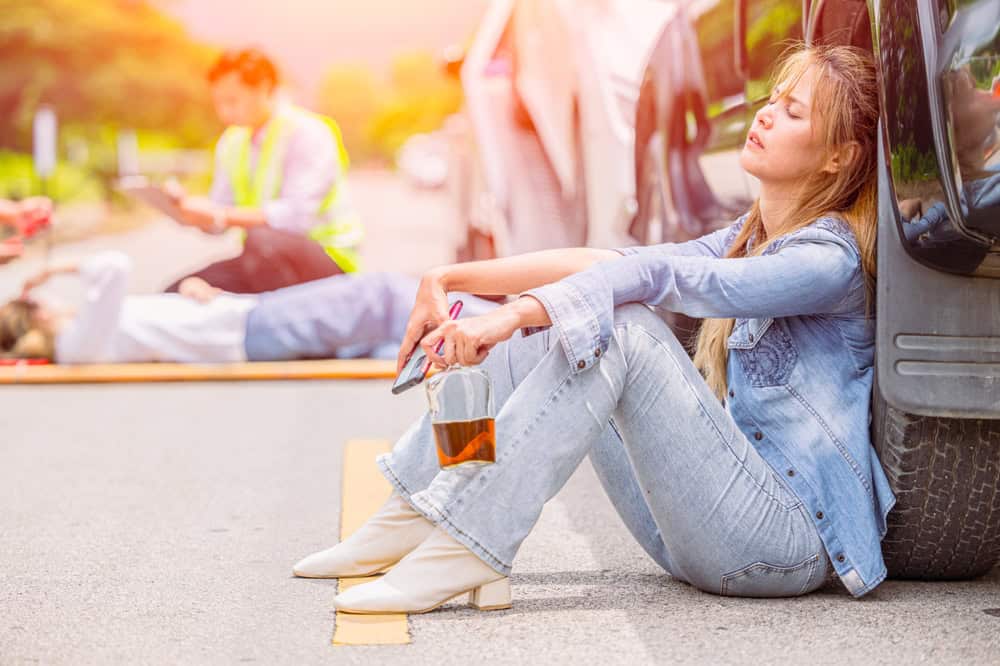 A woman sitting on the roadside, visibly distressed, near a car involved in an accident. Emergency responders are nearby.