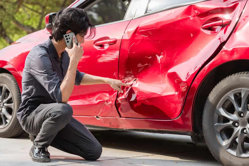 A man stands next to a red car, talking on his cell phone, possibly after a car accident.