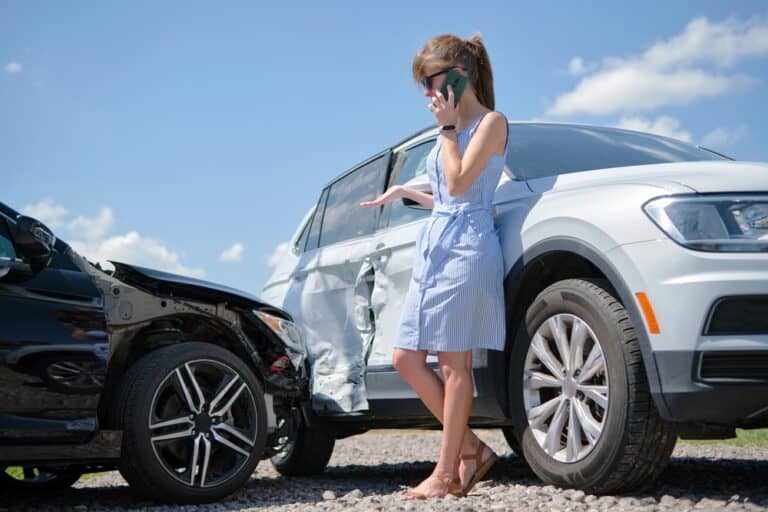 A woman on her cell phone next to a damaged car, calling 911 after a car crash.