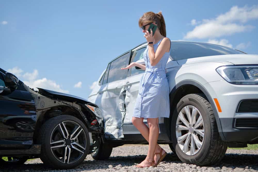 A woman on her cell phone next to a damaged car, calling 911 after a car crash.